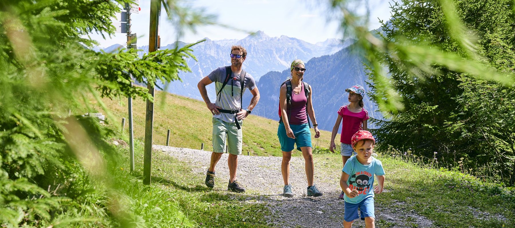Familie wandert im Sommer auf Waldweg mit Blick auf Berge und grüne Almwiesen