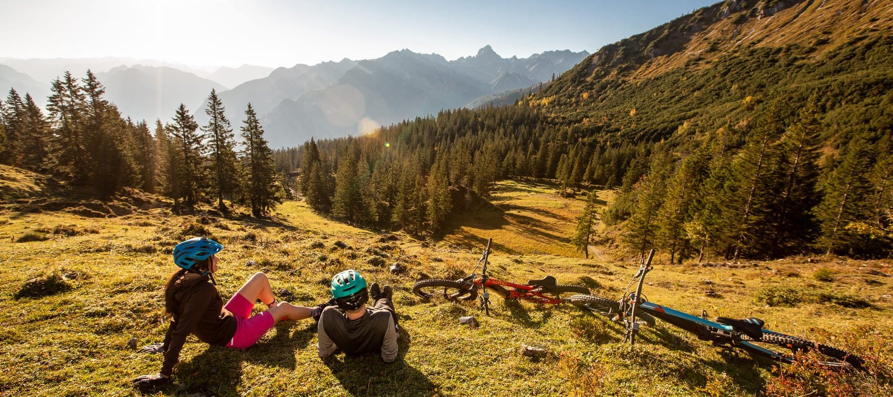 Zwei Mountainbiker liegen auf einer Almwiese und erholen sich bei strahlendem Sonnenschein mit Alpenpanorama