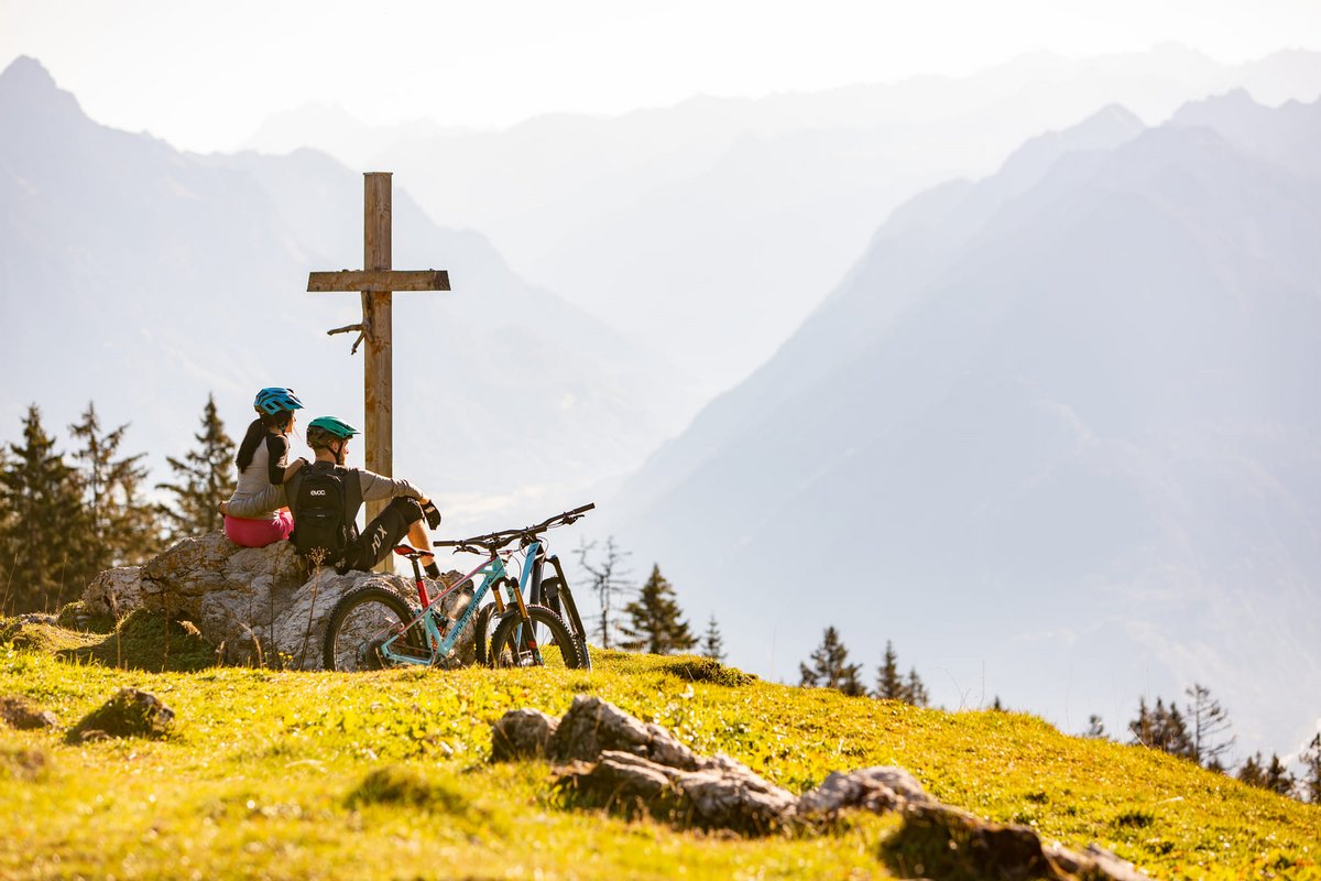 Zwei Mountainbiker rasten bei einem Gipfelkreuz und genießen den Ausblick auf die Alpen im Brandnertal