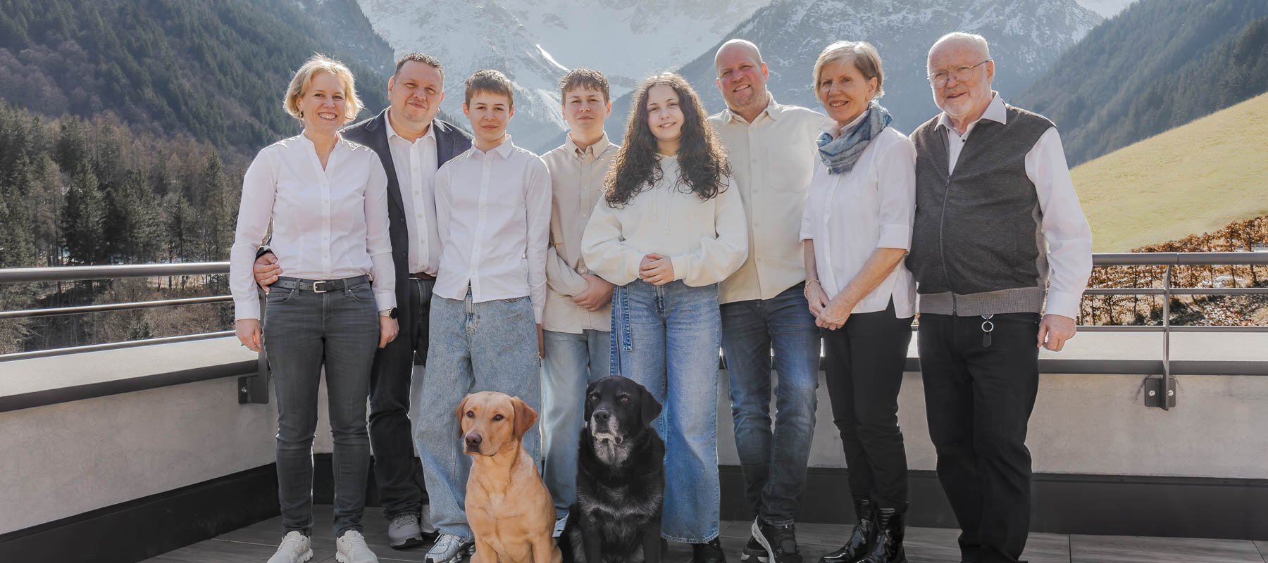 Mehrgenerationen-Familie mit zwei Hunden auf einer Terrasse vor alpiner Bergkulisse